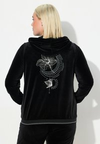 Chaqueta con capucha de terciopelo negro que presenta un diseño gráfico blanco de flores y patrones circulares en la parte trasera, con un ribete en contraste en el dobladillo.