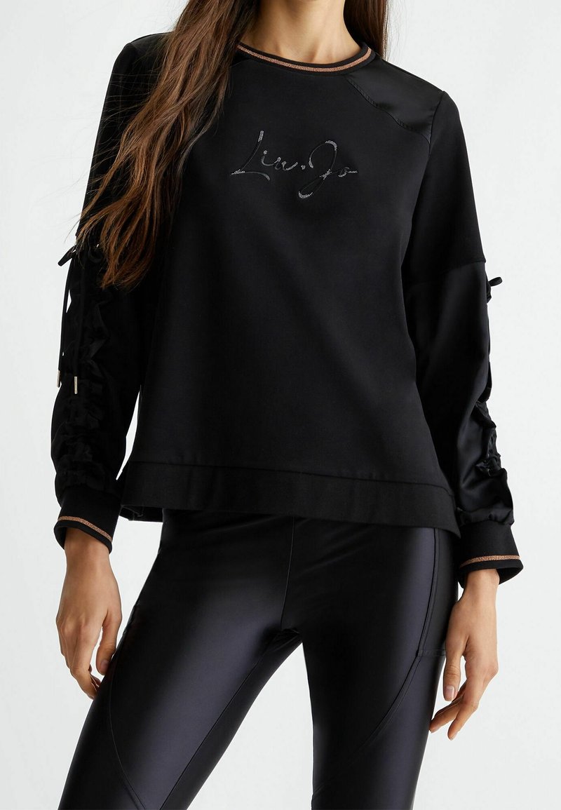 Femme portant un haut noir à manches longues avec un texte en cursive et des leggings noirs brillants et ajustés, debout devant un fond uni.