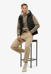 Czarna kamizelka puchowa noszona na beżowym hoodie, w połączeniu z beżowymi spodniami i jasnymi trampkami. Outfitu dopełniają podwinięte mankiety i wzorzyste skarpetki.