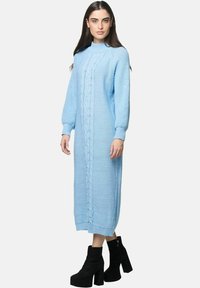 Kvinde iført en lyseblå lang strikket sweaterkjole med kabelmønster og sorte platform ankelstøvler, stående foran en ensfarvet baggrund.