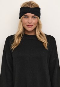 Diadema negra de punto con un diseño retorcido, llevada con un suéter negro holgado. El material parece suave y texturizado, creando un aspecto acogedor.