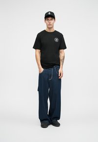 Svart bomull T-skjorte med rund halslinning og logo-trykk, kombinert med mørkeblå stripete vide bukser og svarte sneakers.