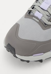 Chaussure de sport gris clair avec tige en mesh, semelle en caoutchouc gris foncé, motifs texturés et accents violets sur les lacets et le côté.