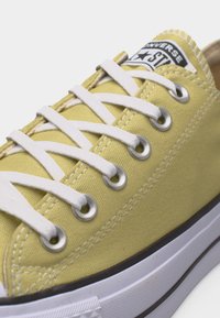 Κίτρινο πάνινο παπούτσι Converse με λευκά κορδόνια, μεταλλικά οπές και λευκή λαστιχένια σόλα με μαύρη ρίγα.