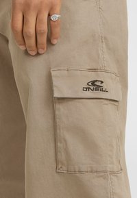 Vaaleat beige cargo-housut sivutaskulla, jossa on tumma brodeerattu logo. Silkkinen puuvillakangas ja suorasaumainen muotoilu.