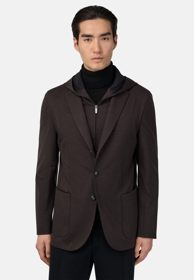 Tumši brūns pielāgotais žakets ar kapuci, featuring divām priekšējām kabatām, diviem pogām un gludu tekstūru. Valkāts virs melna turtleneck.