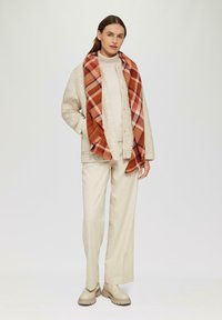 Beige wollen jas, lichte trui, beige broek en chunky beige laarzen. Gestreepte sjaal met een rood, oranje en roze ruitpatroon.