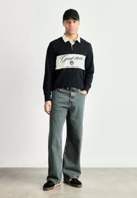 Polo cu maneci lungi de culoare bleumarin și crem, cu un logo și text, asortat cu blugi gri largi și adidași negri cu talpă bej.