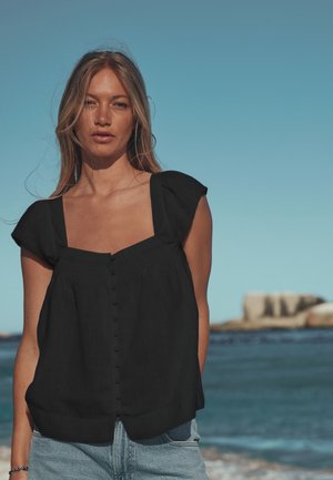 Jeune femme aux longs cheveux blonds portant un haut noir sans manches et un jean bleu clair, debout près d'une mer calme sous un ciel bleu clair.