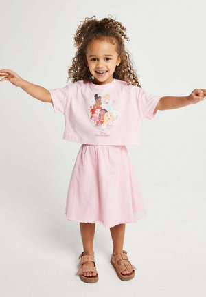 Lächendes junges Mädchen mit lockigem Haar trägt ein rosafarbenes Disney-Prinzessinnen-Oberteil und einen Rock, steht mit ausgestreckten Armen vor weißem Hintergrund.