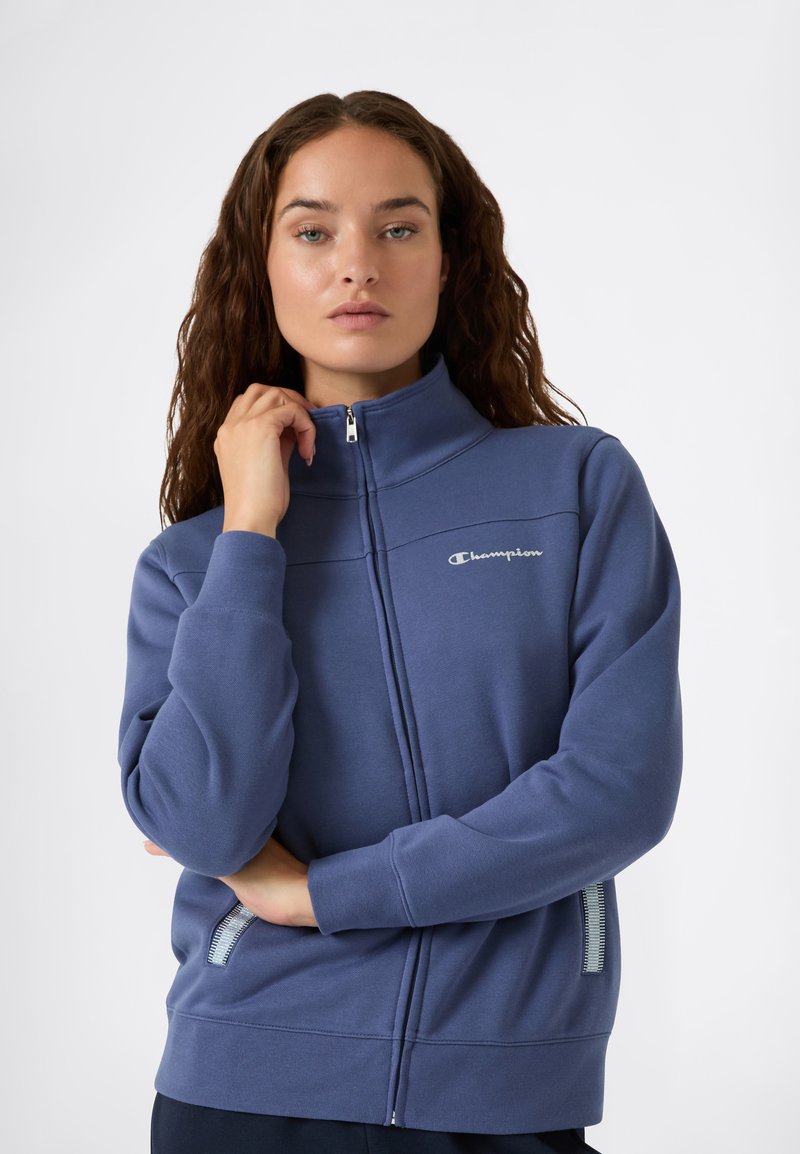 Felpa con zip di un blu scuro, dotata di un alto colletto, logo Champion sul petto e dettagli testurizzati sulle tasche laterali.