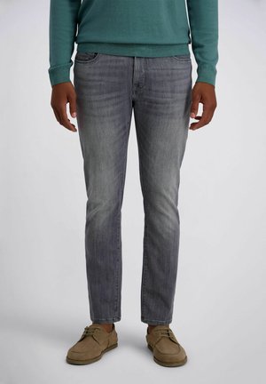 Mann trägt graue Slim-Fit-Jeans, grünen Langarmpullover und braune Schnürschuhe, steht vor einem einfarbigen grauen Hintergrund.