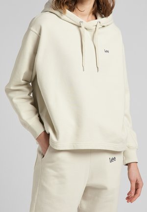 Femme portant un sweat à capuche crème de la marque Lee et un pantalon de survêtement assorti, une main dans la poche sur fond uni.