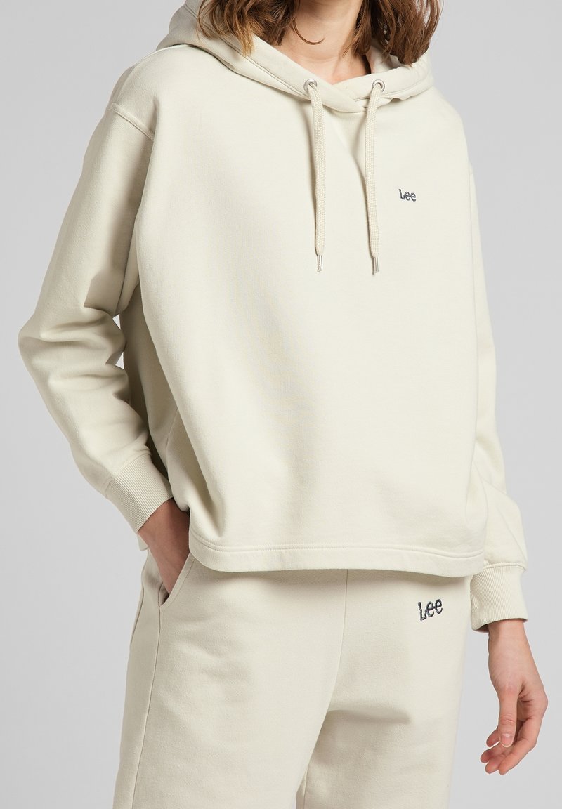 Femme portant un sweat à capuche crème de la marque Lee et un pantalon de survêtement assorti, une main dans la poche sur fond uni.