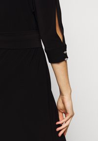 Ruka ženy v černé šaty s rukávem a otevřeným rozparkem, se dvěma ozdobnými stříbrnými knoflíky poblíž zápěstí na jednoduchém pozadí.