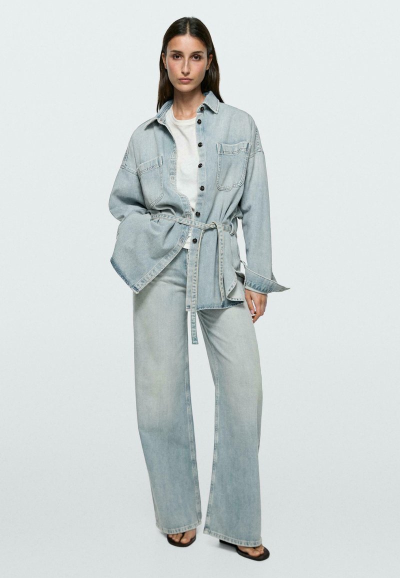 Vrouw staand, draagt een lichtblauwe denimjas met ceintuur over een wit shirt, gecombineerd met lichtblauwe denim jeans met rechte wijde pijpen en zwarte open schoenen.