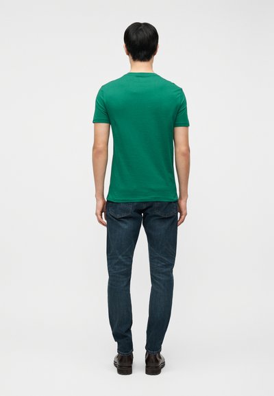 Άνδρας που στέκεται με την πλάτη στραμμένη, φοράει πράσινο κοντομάνικο πουκάμισο, σκούρο μπλε τζιν και σκούρα καφέ παπούτσια, σε απλό φωτεινό φόντο.