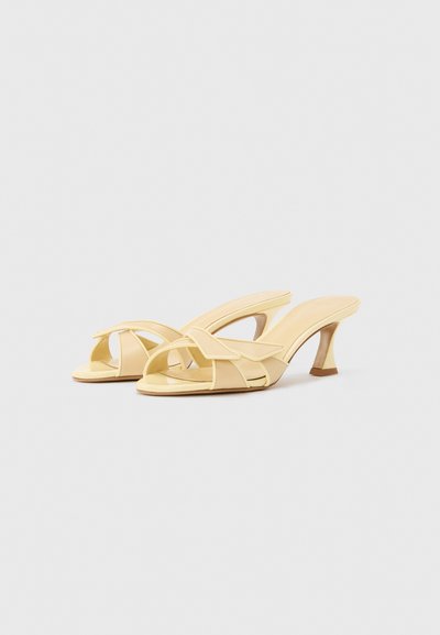Ζευγάρι παστέλ κίτρινων σανδαλιών με τακούνια και ανοιχτά δάχτυλα, με σταυρωτά λουράκια, σχεδιασμένα με καμπυλωτά τακούνια μέσου ύψους σε απλό φόντο.