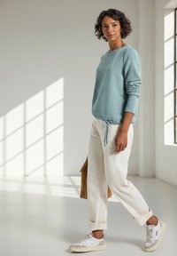 Femme marchant à l'intérieur, portant un sweat-shirt bleu clair, un pantalon blanc retroussé, des baskets blanches et portant un sac marron.