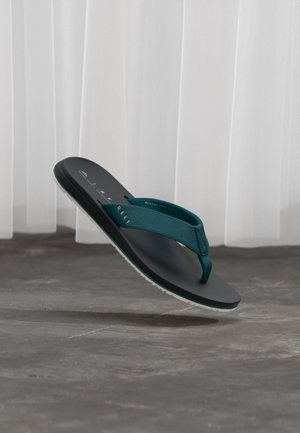 Zwarte sandaal met turkooizen bandjes zwevend boven een grijze vloer met witte gordijnen op de achtergrond.