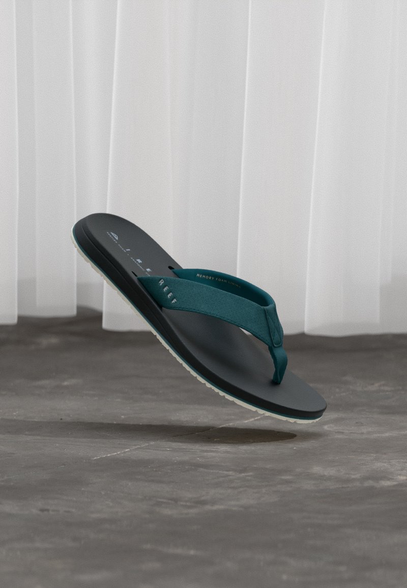 Μαύρο σανδάλι με τιρκουάζ λουράκια που αιωρείται πάνω από γκρι δάπεδο με λευκές κουρτίνες στο φόντο.