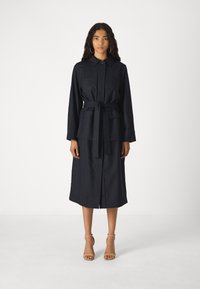 Navy pinstriped dress with a collar, front-button closure, tie waist, and two chest pockets, reaching mid-calf. Worn with nude heels.