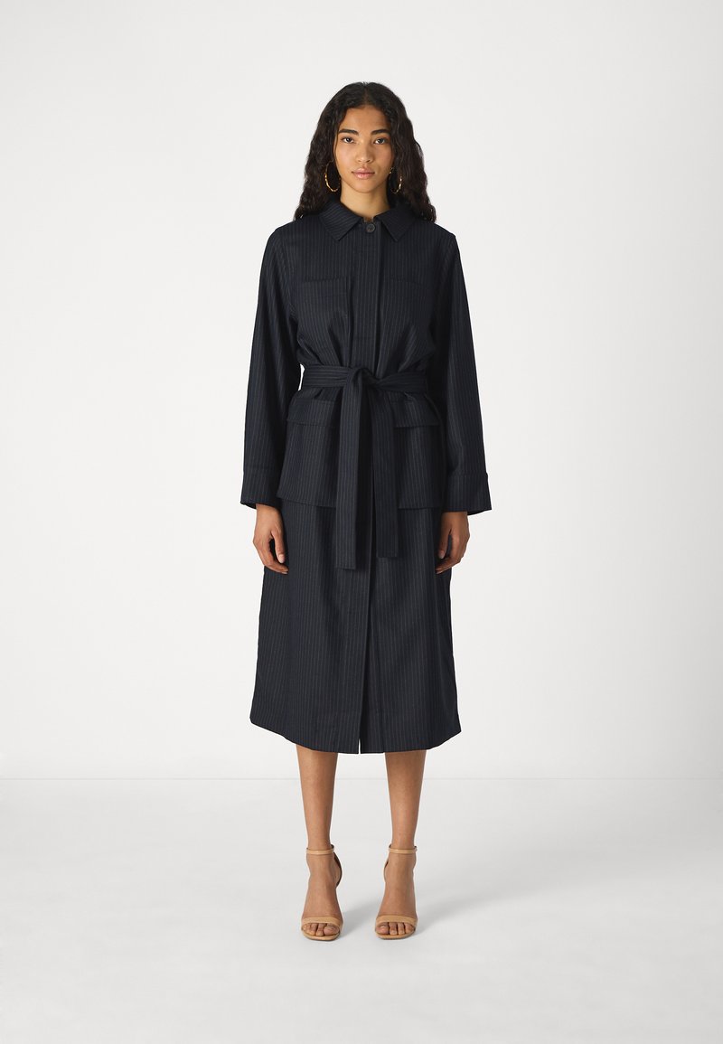 Navy pinstriped dress with a collar, front-button closure, tie waist, and two chest pockets, reaching mid-calf. Worn with nude heels.