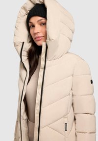 Beige puffer jacket with a large hood, quilted design, zip closure, and black accent details. Worn over a light-colored top.