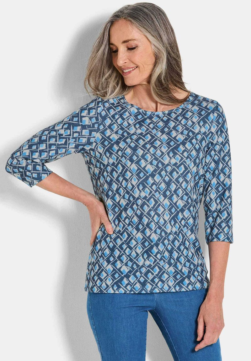 Blau gemusterte Bluse mit Dreiviertelärmeln, die ein geometrisches Design in verschiedenen Blau- und Weißtönen aufweist. Weicher Stoff.