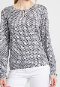 Langärmliges Oberteil in einem schwarz-weißen geometrischen Muster, mit einem Schlüsselloch-Ausschnitt und strukturierter Stoffqualität. Kombiniert mit weißen Hosen.