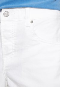 Jean en denim blanc avec une texture lisse, mettant en avant des accents de boutons métalliques, des détails de poches cousus et une coupe ajustée.