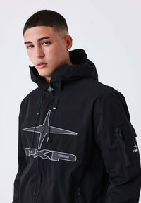 Giovane con capelli corti che indossa una giacca nera con cappuccio, caratterizzata da un grande logo bianco a forma di stella e tasche con zip sul petto e sulla manica.