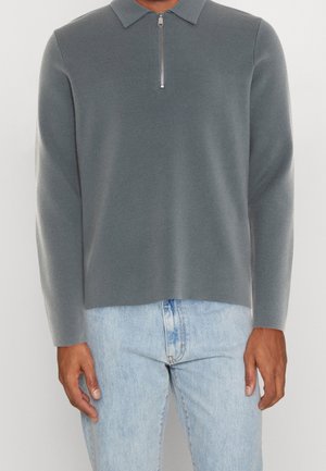 Man draagt een grijze pullover met lange mouwen, een korte ritssluiting aan de voorkant en lichtblauwe spijkerbroek.
