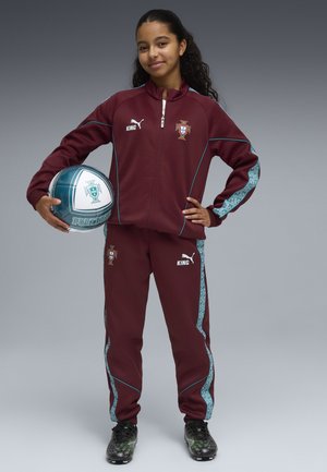 Ung pige i vinrød Portugal træningsdragt, der holder en fodbold med Portugal-emblem, stående med den ene hånd på hoften.