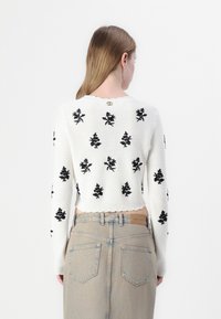 Maglione corto lavorato a maglia bianco con ricami floreali neri, polsini e orlo a costine, e un bottone decorativo sul retro. Indossato con pantaloni in denim chiaro.