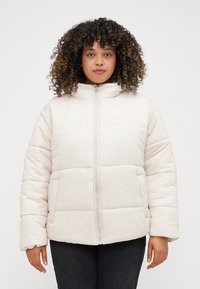 Ανοιχτό μπεζ μπουφάν τύπου puffer με ψηλό γιακά, φερμουάρ μπροστά και δύο πλαϊνές τσέπες. Διαθέτει ραφές τετράγωνων τμημάτων και ένα μικρό λογότυπο στο στήθος.