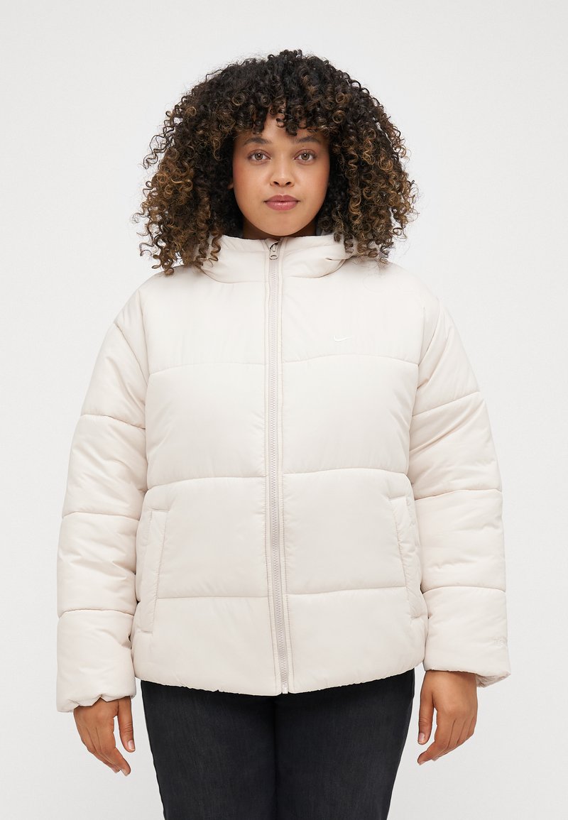 Ανοιχτό μπεζ μπουφάν τύπου puffer με ψηλό γιακά, φερμουάρ μπροστά και δύο πλαϊνές τσέπες. Διαθέτει ραφές τετράγωνων τμημάτων και ένα μικρό λογότυπο στο στήθος.