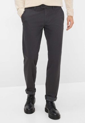 Chinos grigio scuro in misto cotone, con gamba dritta e orli arrotolati. Presentano tasche anteriori e chiusura con bottone. Stivali neri in pelle indossati.