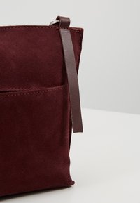 Vínově červená semišová taška s hnědým koženým páskem a předním kapsou. Texturovaný povrch s dekorativním prošitím podél okrajů.