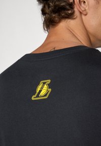 Sort bomulds T-shirt med et gult logo med en basketball og bogstavet "L" på ryggen, korte ærmer og rund halsudskæring.