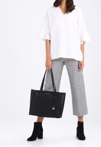 Sac fourre-tout en cuir noir à finition texturée, avec double poignée et accent métallique. Porté avec un haut blanc, un pantalon rayé et des bottines noires.