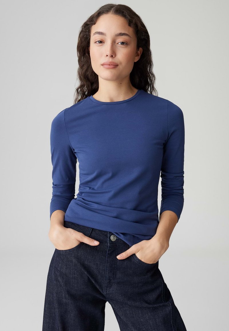 Königsblaues Langarmshirt mit rundem Ausschnitt, glatter Textur und figurbetontem Schnitt. Kombiniert mit dunklen Denimhosen mit Taschen.