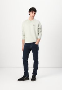 Felpa girocollo verde chiaro con un piccolo logo a patch, abbinata a jeans blu scuro e sneakers nere. Sfondo semplice, modello in posa con aria di sicurezza.