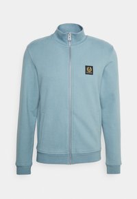 Sudadera azul con cremallera y cuello alto, hecha de un material suave. Cuenta con un pequeño parche negro en el pecho, bolsillos laterales y puños acanalados.