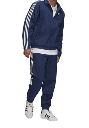 Donkerblauwe trainingspak van lichtgewicht stof met witte drie-strepen accenten op de mouwen van de jas en de broek, gecombineerd met klassieke witte sneakers.