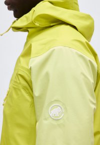 Homme portant une veste à capuche jaune-vert avec un logo de mammouth blanc brodé sur le haut du bras.