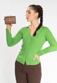 Cardigan verde a coste con colletto, dotato di chiusura a bottoni. Tiene in mano un portafoglio marrone con texture e lettering dorato, posizionato su uno sfondo bianco.