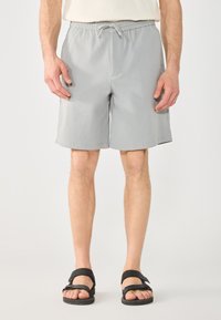 Shorts de bain gris clair avec une taille élastique, un cordon de serrage et des poches latérales, portés par une personne en sandales noires.