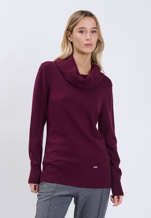 Vrouw die een paarse geribbelde coltrui en grijze broek met kleine witte stippen draagt, staand tegen een effen lichte achtergrond.