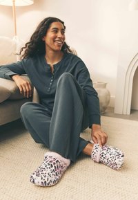 Hyggelige leopardmønstrede hjemmesko med lyserød pelsfor, båret med mørkegrå loungewear, på et beige tekstureret tæppe ved siden af en lys sofa.